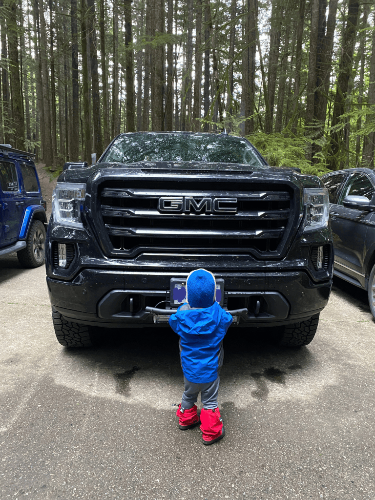A child stands in front of a parked pickup truck. The child's head reaches barely above the license plate of the truck. 
